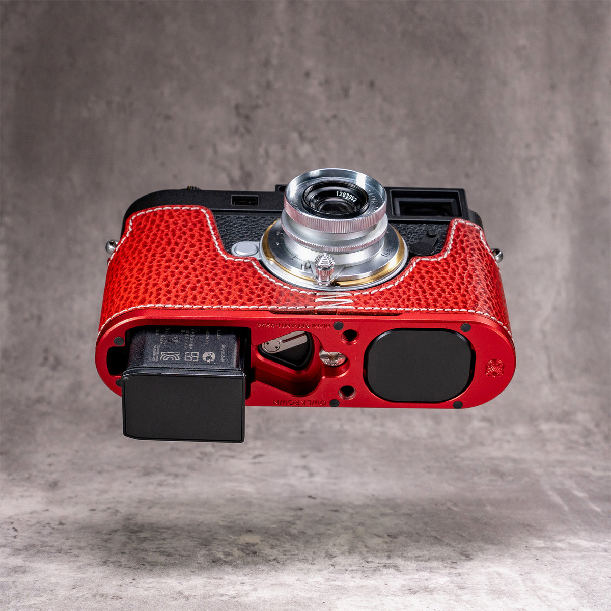 Battery released from a Leica M11 with the Ruby Gloss FlexCase still fitted, AirTag cover cap in place on the OxBlood plate