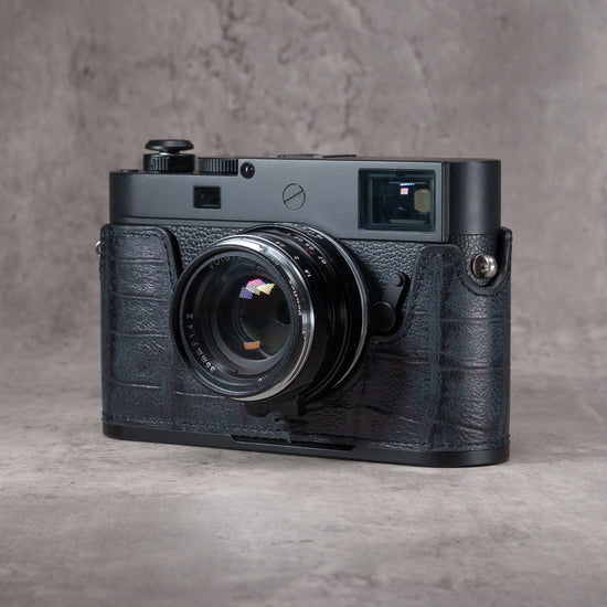 Leica M camera in a Black Swamp gator pattern FlexCase with a Black Chrome plate, front three-quarter view from the left on a grey studio backdrop