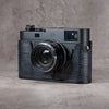 Leica M camera in a Black Swamp gator pattern FlexCase with a Black Chrome plate, front three-quarter view from the left on a grey studio backdrop