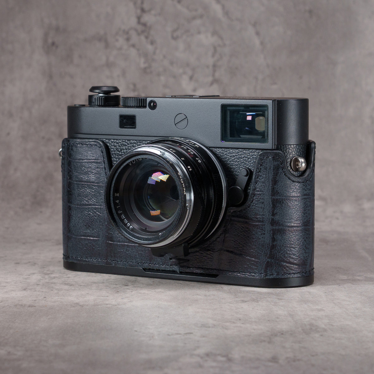 Leica M camera in a Black Swamp gator pattern FlexCase with a Black Chrome plate, front three-quarter view from the left on a grey studio backdrop