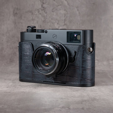Leica M camera in a Black Swamp gator pattern FlexCase with a Black Chrome plate, front three-quarter view from the left on a grey studio backdrop