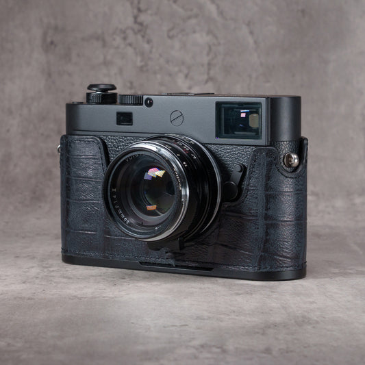 Leica M camera in a Black Swamp gator pattern FlexCase with a Black Chrome plate, front three-quarter view from the left on a grey studio backdrop